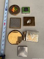 Photo of seven assorted vintage makeup compacts arranged beside a measuring tape for scale, showing different shapes, sizes, and designs.
