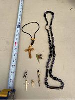 Image shows a measuring tape for scale alongside seven jewelry items on a flat surface: a black beaded necklace with a wooden cross, a long purple and black beaded necklace, a colorful hummingbird brooch, two small gold-tone cross earrings, and two gold-tone pins including one with a pearl-like bead.