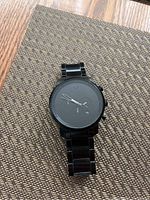 Photo showing an all black MVMT wristwatch with a metal strap, placed on a textured surface.
