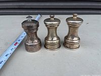 Three vintage silver plated pepper mills arranged in a row on a surface, with a measuring tape alongside for scale.