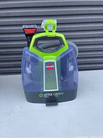 Photo of a Bissell Little Green ProHeat Pet carpet cleaner showing the front with transparent water tanks, hose attachment, and handle.