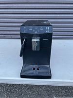 Front view of black Saeco espresso machine on a white surface against a corrugated metal backdrop. Shows control buttons, steam wand, drip tray, and brand name.