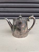Photo of silver tone single serve tea pot with curved spout, decorative handle, and knob lid showing some tarnish and unusual surface wear.
