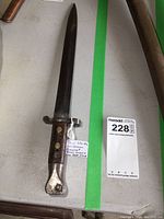 Full view of antique Wilkinson bayonet showing blade and handle with aged wear and attached tag.