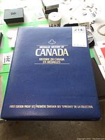 Closed blue presentation book titled 'Medallic History of Canada First Edition Proof Set' with gold and silver embossing.
