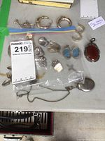 Photo of various silver jewelry including crescent-shaped pins/brooches, cufflinks, several small brooches or pins in plastic bag, a reddish stone pendant, and a silver locket with chain.