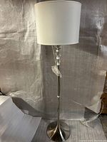 Full view of one floor lamp showing white cylindrical fabric shade, metal base, and clear glass accents on the pole.