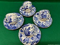 Four Royal Crown Derby Mikado tea cups and saucers arranged on a green cloth, showing intricate blue and white Asian-themed decorations.