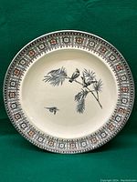 Front view of an antique decorative plate with black and orange geometric borders and central bird and branch design on a cream background.