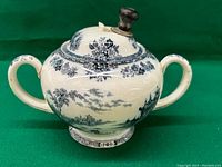 Front view of antique Royal Doulton ceramic bowl with lid, cream-colored with black scenic transferware, showing round bulbous body, two handles, and knob on lid.