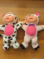 Two Cabbage Patch Kids dolls, one in a black and white cow costume with pink belly, the other in a pale pink pig costume with bow tie, displayed laying flat on a wooden surface.