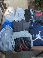 Overview photo showing the full lot of kids clothes including various sweatshirts, sweatpants, shorts, and jackets from brands Adidas, Puma, Jordan, and Under Armour.