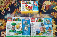 Photo showing all three Lego Super Mario sets in sealed packaging arranged on a patterned carpet, highlighting the set numbers and character images.