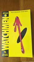 Front cover of Watchmen graphic novel showing yellow background with stylized blood-stained smiley face design and text.