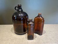 Three amber/brown vintage antique bottles displayed on a countertop: a large jug, a medium curved whiskey bottle, and a small rectangular embalming fluid bottle.