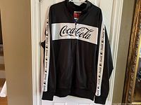 Front view of the black and white Coca-Cola zip-up jacket with logo on chest and sleeves.