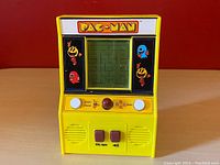 Front view of yellow Pac-Man handheld game showing screen with maze and character graphics, control buttons, and power switches.