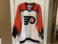 Front view of the Philadelphia Flyers white, orange, and black hockey jersey hanging on a hanger showing the front logo.