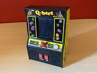 Front angled view of the yellow and black Q*bert handheld video game showing screen, control pad, buttons, and character graphics.