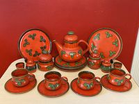 Full view of all 24 pieces of the vintage 70s pottery tea set showing teacups, saucers, plates, teapot, canister, sugar dish, and egg cups arranged in front of a red background.