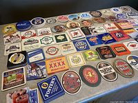 Top view of a table covered with about 50 assorted vintage beer and soda coasters laid out in rows, showing various company logos and colors.