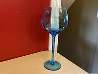 Front angle view of large vintage blue hand blown glass goblet vase with twisted stem on beige surface against a red and beige background.