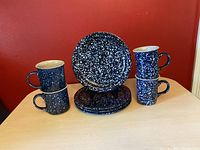 Four blue speckle ceramic items displayed against a red backdrop: two plates standing vertically and two pairs of mugs stacked and placed beside the plates.