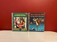 Two vintage children's Christmas Little Golden Books displayed upright against a red background, visible covers for 'The Night Before Christmas' and 'Rudolph the Red-Nosed Reindeer'.