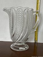 Full side view of the clear glass pitcher showing scalloped top edge and textured vertical panels with handle, measuring about 8.5 inches tall.