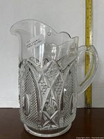Full side view of clear pressed glass pitcher showing intricate diamond and starburst pressed patterns and scalloped rim. An 8 inch measuring tape is visible for scale.