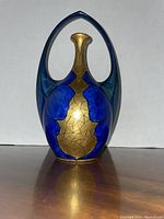 Full view of the Art Nouveau vase with cobalt blue and gold floral/leaf pattern and arching handle, standing on plain light background.