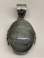 Front view of oval sterling silver pendant with a polished neutral stone with shimmering tones. Intricate silver scrollwork is visible on bail and sides.