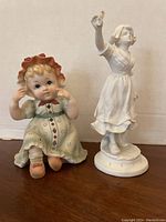 Two antique bisque porcelain figurines displayed side by side on a wooden surface against a neutral background. The left figurine is the Piano Baby figure in pastel colors; the right is the white Sevres style figure.