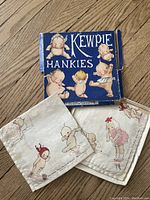 Two printed Kewpie doll handkerchiefs laid out beside their original blue box, showing box wear and handkerchief artwork.