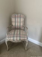 Photo showing a single antique style chair with carved wood frame and plaid upholstery, positioned against an off-white wall on carpeted floor.