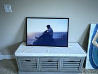 Framed poster laid on top of a bench showing a young man looking out at the sea at sunset, black frame, neutral wall background