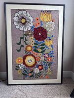 Framed textile art with colorful knitted flowers against beige background in simple black frame.