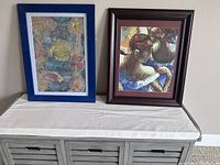 Two framed paintings by Catalina on a white cloth over a gray cabinet. Left painting is underwater fish scene with blue frame, right is a portrait in dark wood frame.