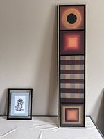 Photo showing one vertical tall frame holding four abstract prints and one smaller black framed print on table next to it.