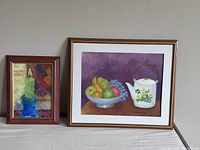 Photo showing two framed paintings set against a wall: a smaller abstract painting to the left and a larger still life painting to the right.