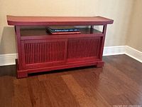 Red Chinese style wooden console with slatted sliding doors and an open shelf, placed against a beige wall on hardwood flooring.