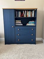 Front view of the blue dresser showing three drawers with wooden knobs, two open shelves above with books, and a cupboard door on the left. The dresser has a natural wood top and blue painted body.