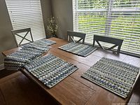 Photo of a wooden dining table with five of the colorful woven placemats arranged flat, showing the texture and colors clearly.
