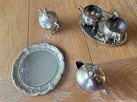 Photo of silver tea set items placed on wood table including tray, creamer, sugar bowl, small two-handled bowl, and decorative plate.