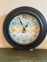 Photo of round wall clock with black frame and colorful chevron pattern on face, mounted on wall.