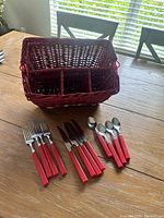 Silverware with red handles arranged in front of a red wicker basket with three divided compartments on a wooden table.
