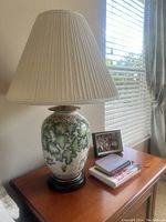 Photo of one night lamp showing ceramic base with green ivy pattern, cream pleated shade, placed on wooden surface next to window and books