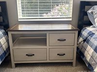 Front view of a wooden night table placed between two beds. The night table has three drawers with black handles and an open shelf on the upper left side. Top surface is a darker wood finish, and body is painted a light color with a distressed look.