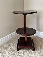 Photo shows a small wooden decorative table with two circular tiers and a square base, dark stained wood finish, placed against a white baseboard corner on carpeted floor.