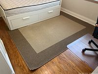 Rectangular beige and brown area rug placed on wooden floor beneath a bed with white drawers and beside a black office chair on a small plastic mat. Shows overall condition and color of rug with visible dirt marks.